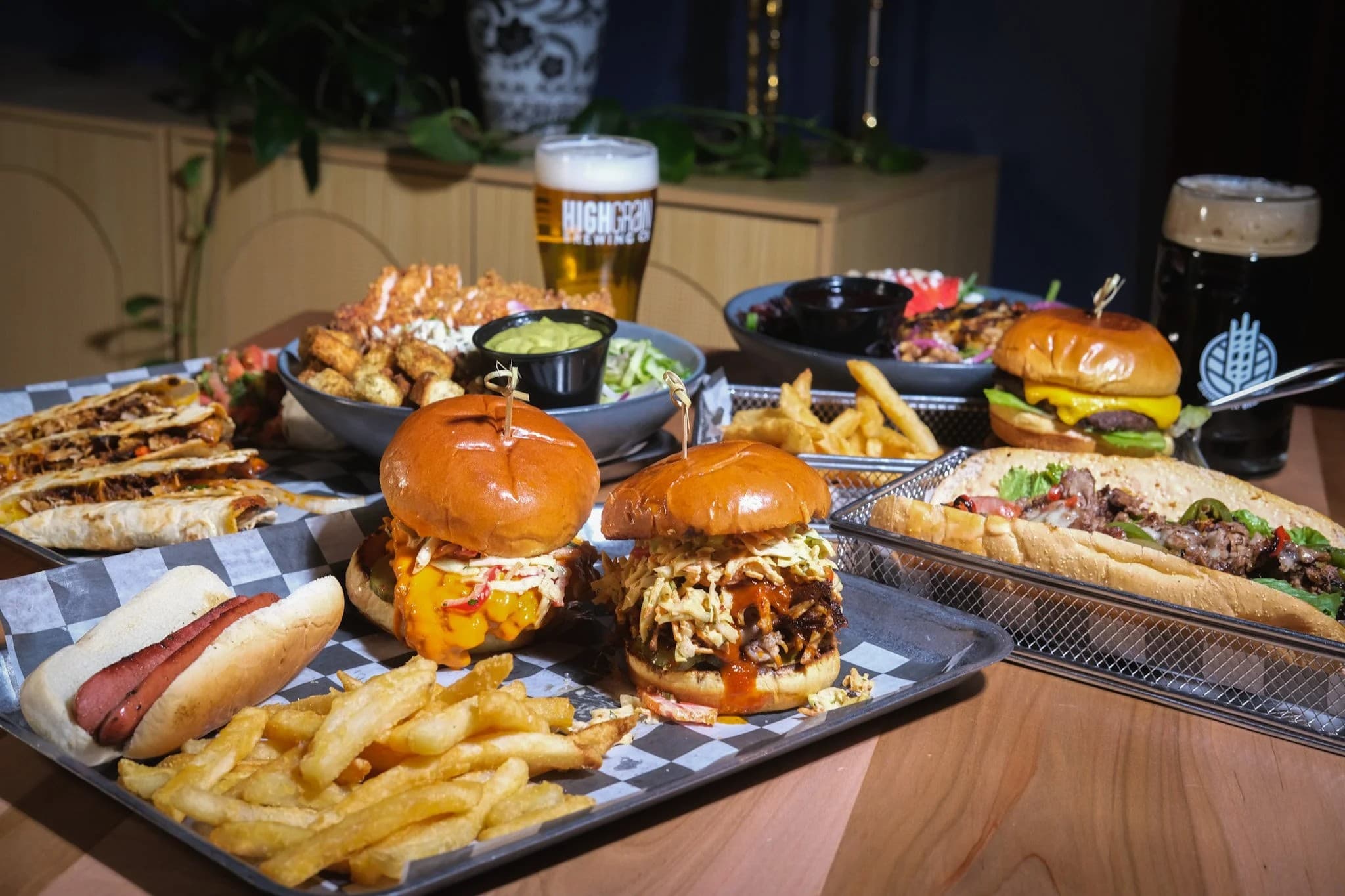A meal consisting of a fried chicken sandwich with a bun, fried chicken, and bacon, a glass of beer with foam on top, and a basket of waffle fries on a wooden table.