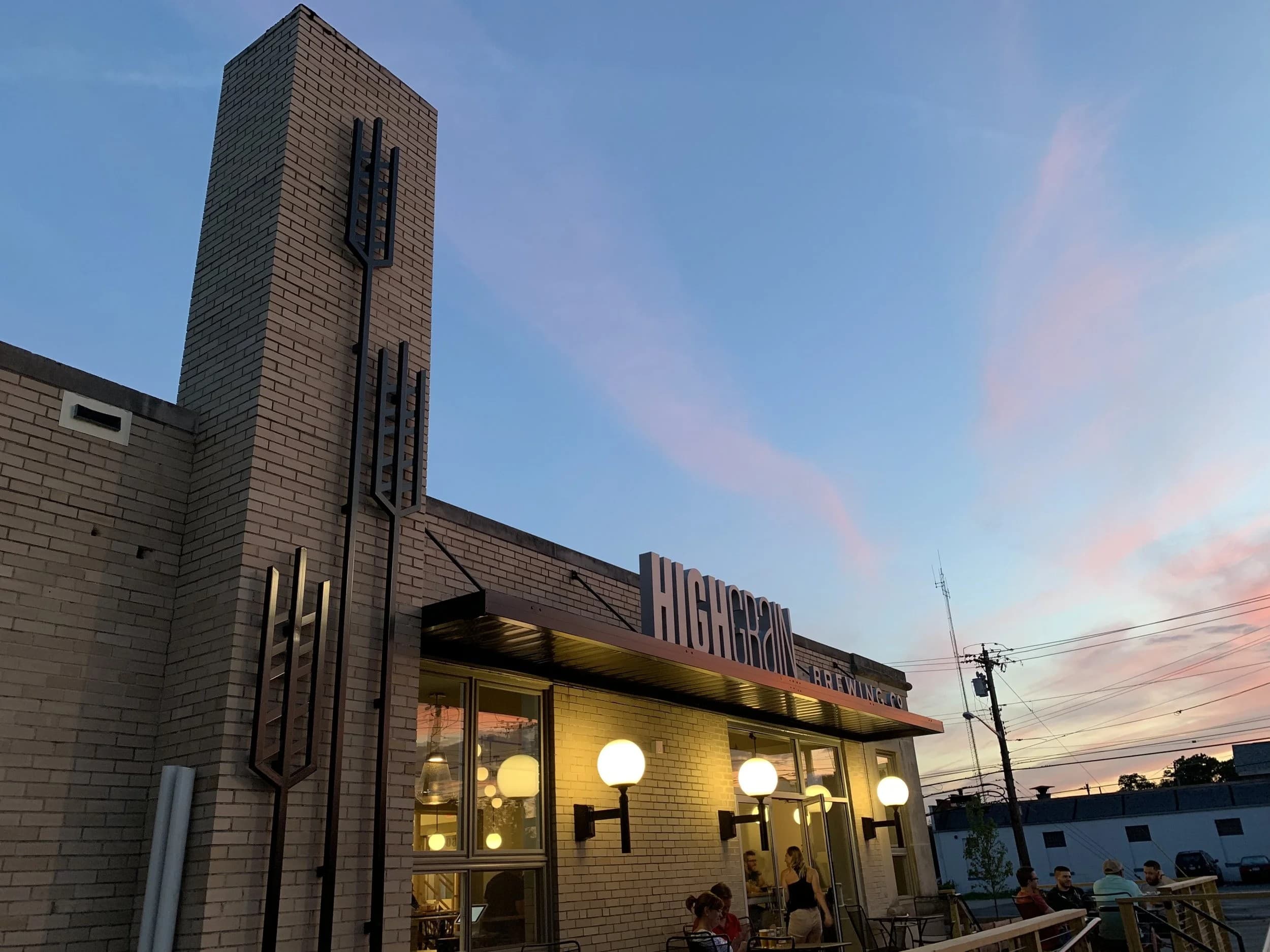 The HighGrain Silverton taproom at sunset: brick tower, decorative metal art, glowing lights and people on the patio.