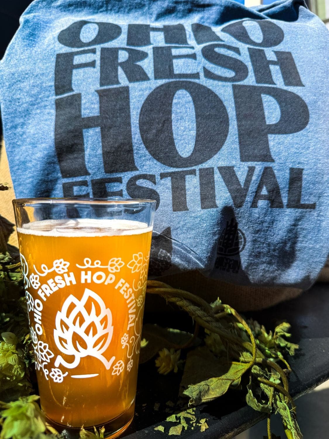 An Ohio Fresh Hop Festival pint glass of hazy beer in front of the festival t-shirt, surrounded by fresh hops