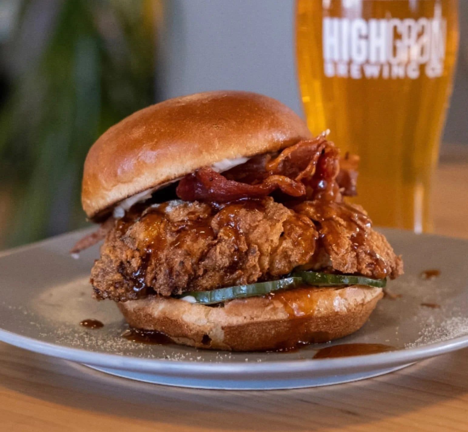 Fried chicken sandwich with pickles, bacon, and a bun on a white plate, with a pint of beer in the background.
