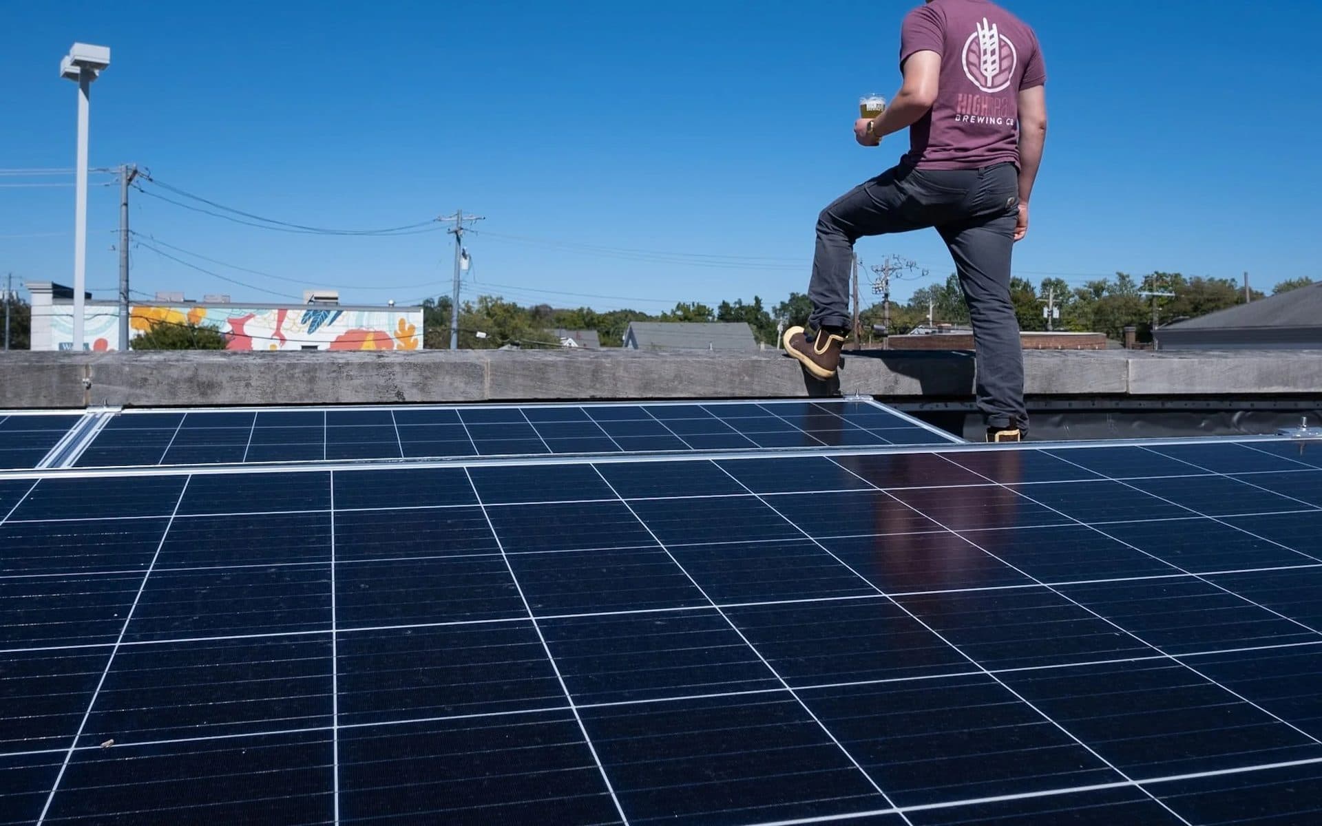 Person standing on a rooftop next to solar panels, holding a glass of beer, under a clear blue sky.