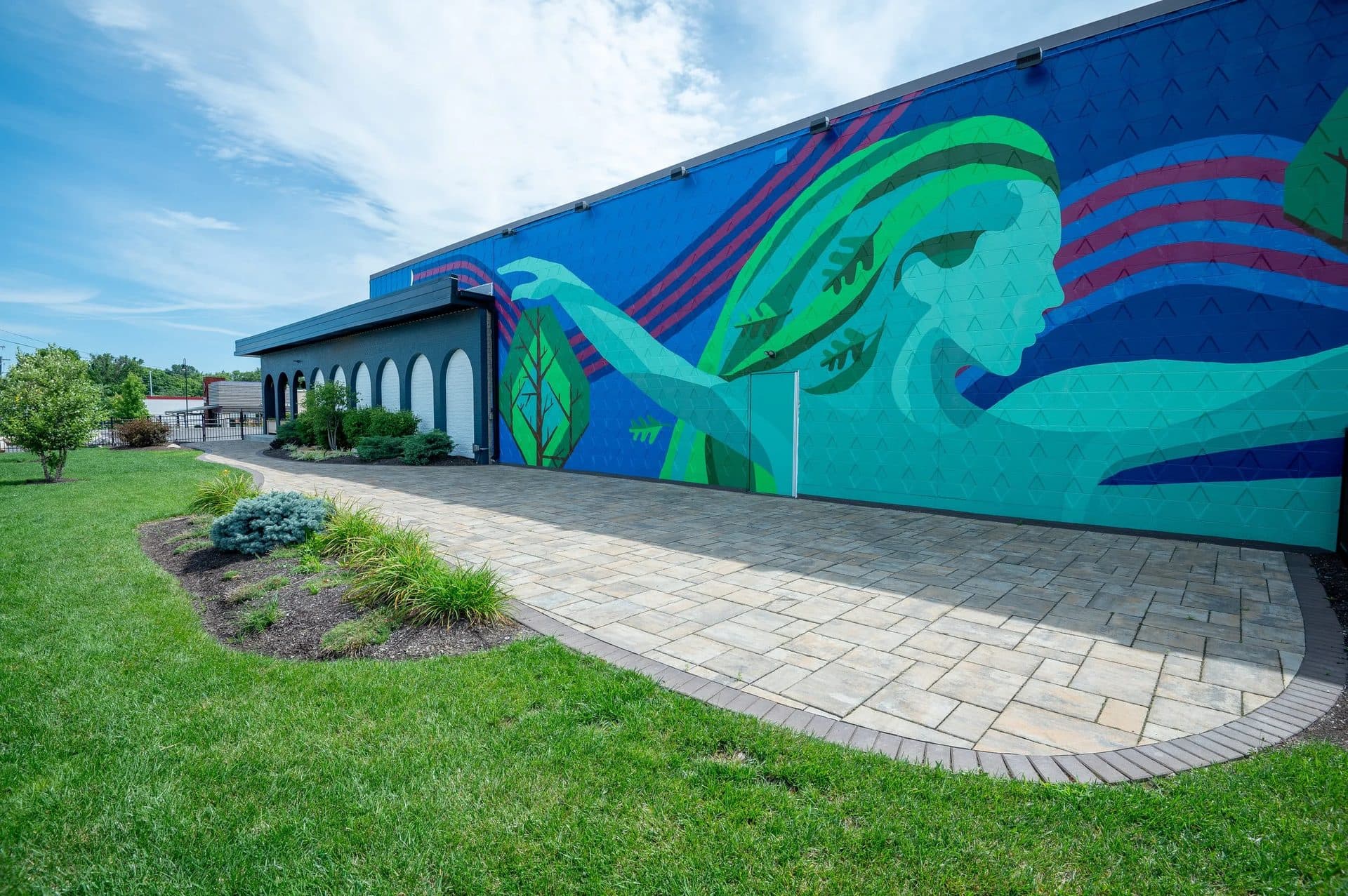 Colorful mural of a woman with long hair reaching out on a building wall, with surrounding trees and a paved walkway in the foreground.