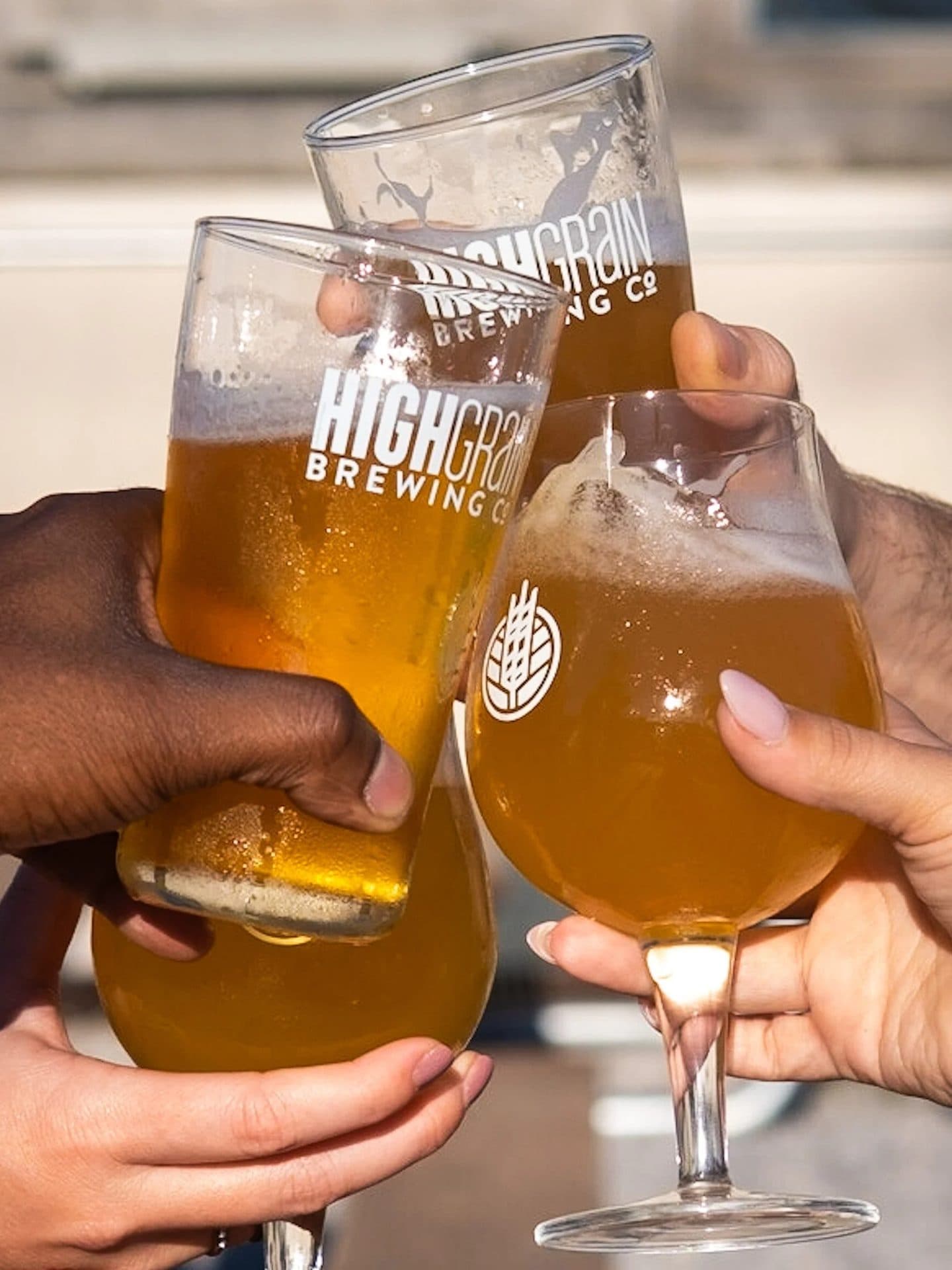 Four hands holding glasses of beer in a toast, with two tall glasses and two stemmed glasses, all filled with golden beer, in front of a blurred outdoor background.